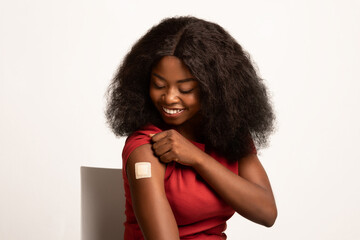 A happy African American woman examines her arm, showing a bandage after receiving her coronavirus vaccine. She poses confidently against a white background, celebrating her health choice.