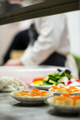 Colorful sushi plates wait as a chef skillfully prepares more ingredients in the background