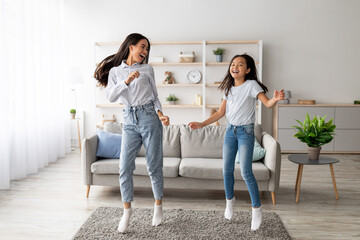 A playful Korean mother and her pretty daughter are dancing and jumping to music in their bright living room. They are enjoying their favorite song and making joyful memories together.