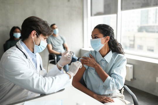 Female patient young brunette woman looking at handsome doctor while getting vaccination against coronavirus at clinic, wearing protective face masks. Immunization while COVID-19 pandemic concept