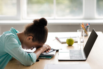 A tired African American teen girl rests her head on the table while surrounded by notebooks and a laptop in her living room. She has just finished an online lesson and feels overwhelmed.