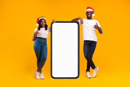 An african american couple stands beside a large cellphone, smiling and giving thumbs-up gestures. They are wearing santa hats and are excited about a holiday offer in a bright yellow studio. - Powered by Adobe