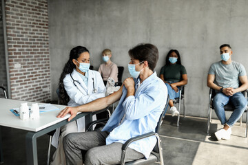 Male patient getting intramuscular shot against COVID-19 at clinic or vaccination center, middle-eastern woman doctor making injection in shoulder for caucasian man, wearing protective face masks