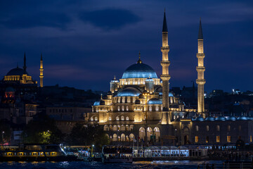 Naklejka premium The New Mosque (Valide Sultan Mosque), beautifully illuminated against the Istanbul night sky, attracts the attention of visitors enjoying the picturesque view of the waterfront.