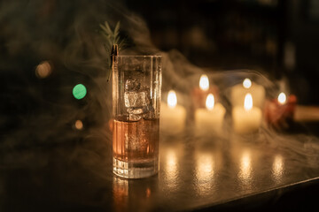 A finished cocktail presented in a dark, moody atmosphere with candlelight and a touch of mist.