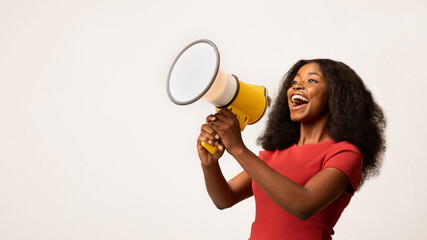 A cheerful African American woman holds a megaphone and smiles while making an announcement. She stands against a white background, showing her excitement and energy.
