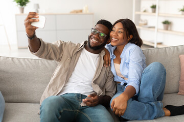 A joyful couple sits on their living room couch, excitedly taking a selfie. The woman leans into the man, both smiling while holding a smartphone. Their relaxed indoor setting adds to the moment.