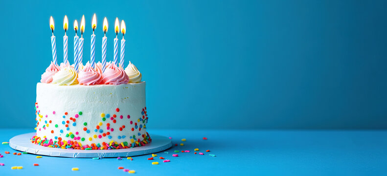 Colorful birthday cake with pink and white frosting, topped with lit candles, surrounded by confetti on a vibrant blue background, perfect for celebration and festive occasions