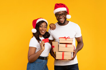 A cheerful African American couple stands together with wrapped Christmas gifts wearing Santa hats. They hold a credit card and smile at the camera, enjoying the festive spirit.