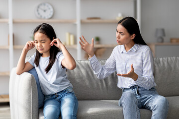In a cozy living room, a young Asian mother speaks to her offended daughter. The girl covers her ears and appears upset, not wanting to listen to her mom's words.