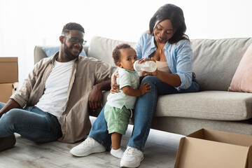 A joyful African American family settles into their new apartment. A loving mother feeds her toddler from a bottle while sitting on the couch, and dad unpacks boxes alongside them.