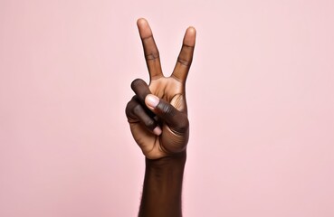 Dark skinned hand shows peace sign gesture. Two fingers up forming V shape. Symbol for victory, peace, or number two. Plain pink backdrop isolates hand.