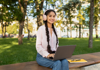 A girl sits on a wooden bench in a green park, using her laptop and wearing headphones. She enjoys the sunny weather surrounded by trees and people in the background.