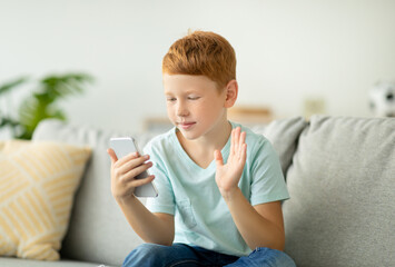 Redhead preteen boy sits on a couch at home, happily waving and smiling during a video call. He is...