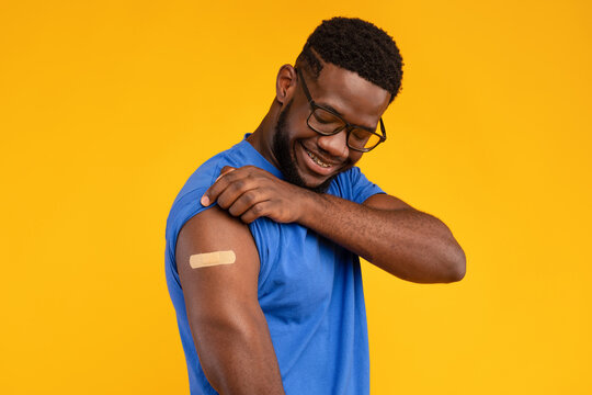 A man smiles as he displays his bandaged arm after receiving the Covid-19 vaccine. The bright yellow background highlights his joyful expression and the importance of vaccination for health.
