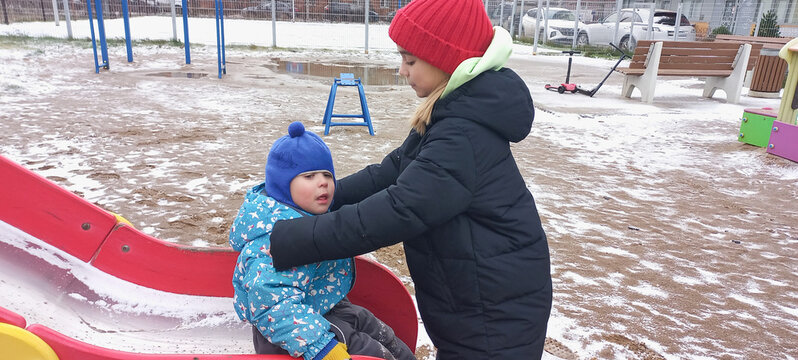 An adult girl helps a child slide down a snowy playground outside