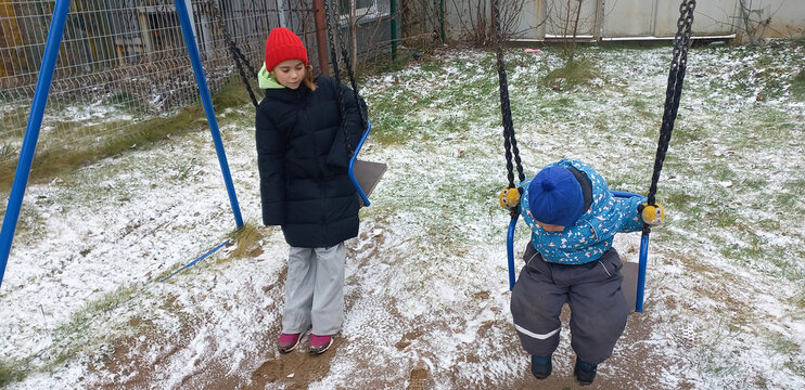 Girl watching boy swing on playground covered in light snow  
