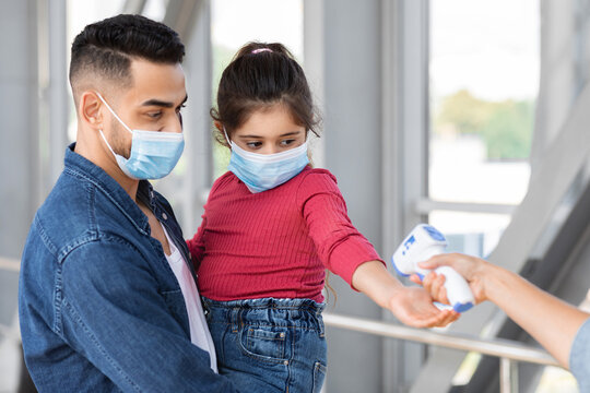 A father holds his daughter as a healthcare worker checks her temperature. Both are wearing masks to follow safety guidelines during the ongoing health situation.