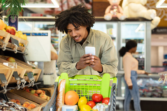 A young man with curly hair smiles as he looks at his phone while pushing a shopping cart filled with fresh fruits and bread in a busy supermarket.