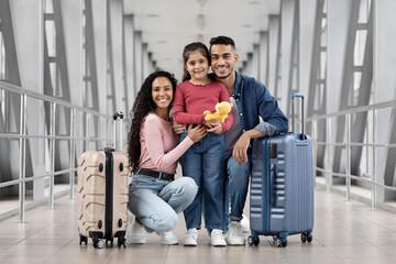 A happy family stands together in an airport terminal, holding a stuffed toy. The parents are...