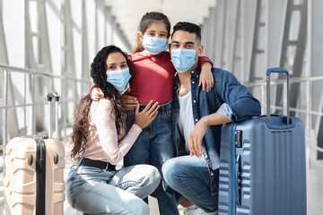 A family of three poses in an airport corridor, wearing face masks for safety. They are smiling while holding their luggage, ready for their journey ahead.
