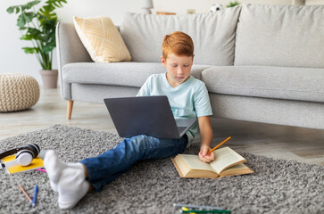 A young ginger boy is focused on his homework at home. He sits on the floor with a laptop and a notebook, looking at a book while writing an essay.