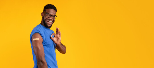 A vaccinated Black man smiles and gestures okay with his hand while showing his arm with a plaster on a bright yellow background. He promotes awareness of vaccination.