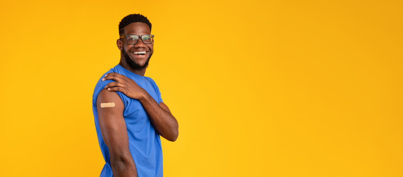 Cheerful African American man smiles while showing his arm with a bandage after getting vaccinated against Covid-19. The bright yellow background enhances the positive atmosphere of the moment. - Powered by Adobe
