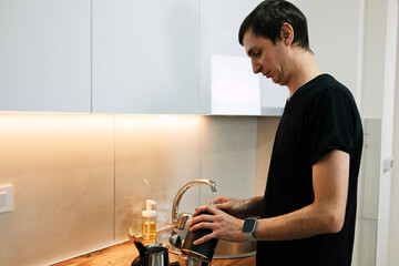 A man holds a coffee maker near the sink while preparing a drink. The scene shows a basic daily ritual and the concept of home comfort.