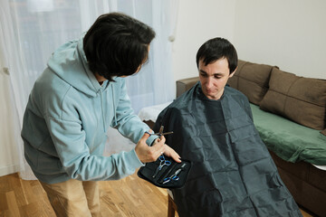 A man sits covered with a haircut cape while woman prepares scissors at home. The scene shows casual grooming and the concept of simple personal care.
