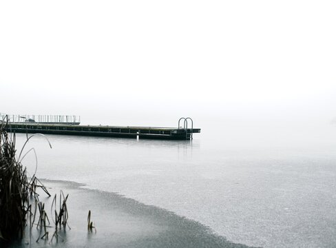 Winter pier in fog A frozen lake with a lone pier and reeds fading into milky fog. Minimalist, cold winter scenery with a calm, muted atmosphere.