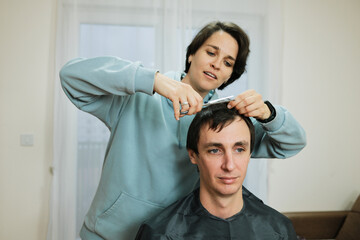 A woman trims a man s hair at home using scissors while he sits calmly. The scene presents everyday grooming and the concept of casual personal care.