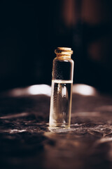 Atmospheric close-up of a small glass vial filled with clear liquid, sealed with a cork stopper, standing on a dark textured surface with warm, moody lighting highlighting droplets inside the bottle.