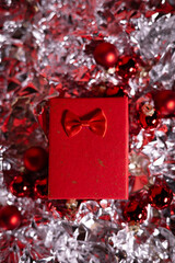 A festive red gift box with a bow surrounded by red Christmas ornaments and delicate white snowflake decorations on a dark green background.