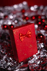 A festive red gift box with a bow surrounded by red Christmas ornaments and delicate white snowflake decorations on a dark green background.