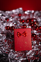 A festive red gift box with a bow surrounded by red Christmas ornaments and delicate white snowflake decorations on a dark green background.