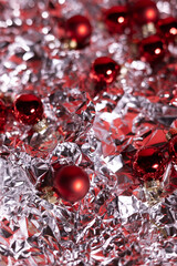 Red Christmas ornaments scattered across a crumpled silver foil surface, creating a festive, reflective, and vibrant holiday background.