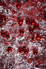 Red Christmas ornaments scattered across a crumpled silver foil surface, creating a festive, reflective, and vibrant holiday background.
