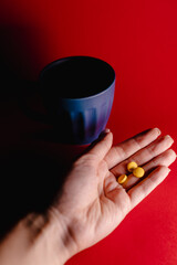Close-up image of a hand holding several yellow tablets against a vivid red background, with a dark blue cup in the background. 