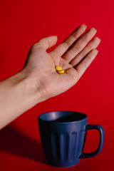 Close-up image of a hand holding several yellow tablets against a vivid red background, with a dark blue cup in the background. 