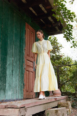 Naklejka premium A woman in a yellow dress stands on porch steps beside a green doorway in a garden setting, enjoying natural daylight and a calm, contemplative moment.
