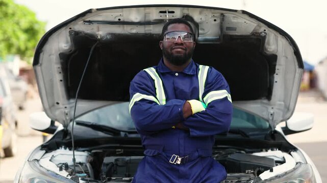 Confident African mechanic, successful small business owner in uniform and safety glasses, standing proudly in front of a car with an open hood at his workshop, SME