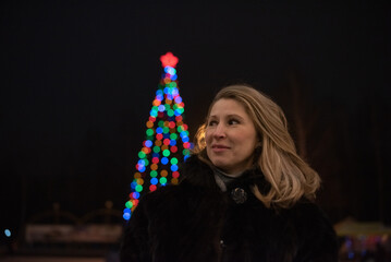Woman with long blonde hair, dressed in a black fur coat, stands outdoors surrounded by colorful...