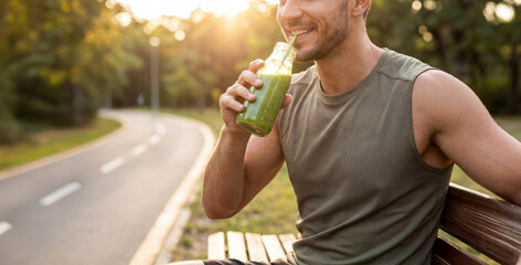 Fitness Man enjoying a refreshing green smoothie drink while sitting on a park bench in the evening sunlight along a winding pathway