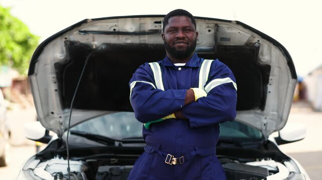 Proud African mechanic, successful small business owner in blue overalls stands confidently in front of a white car with its hood open at his workshop, SME