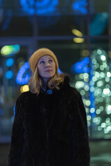 Fototapeta premium Woman with long blonde hair, dressed in a black fur coat, stands outdoors surrounded by colorful holiday lights, creating a festive atmosphere during winter season