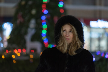 Fototapeta premium Woman with long blonde hair, dressed in a black fur coat, stands outdoors surrounded by colorful holiday lights, creating a festive atmosphere during winter season