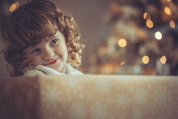 H&uuml;bsches kleines M&auml;dchen mit Locken sitzt in einem Geschenkkarton vor dem Weihnachtsbaum zu Weihnachten, Abendstimmung mit Lichtern Var. 9