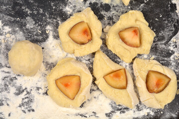 An overhead view of potato dough pieces and plum wedges being prepared on a floured black surface for traditional sweet dumplings