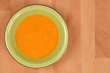 An overhead, close-up shot of a creamy orange carrot and peanut soup served in a green rimmed bowl on a light wooden surface